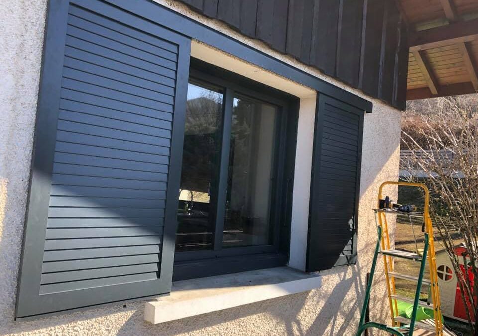 Un mur de maison avec deux volets coulissants ajourés gris anthracite et une menuiserie alu gris anthracite.